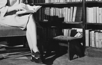A vintage photograph of the legs of a seated person reading a book in front of a large bookcase